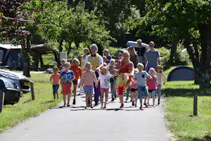 Familie geht auf einem Campingplatzweg zwischen Bäumen, neben geparkten Autos und Zelten.