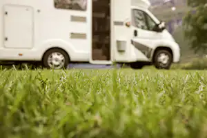 Weißes Wohnmobil steht auf grünem Campingplatz, im Vordergrund Gras, im Hintergrund Hügel und Bäume.