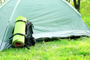 Zelt auf grünem Gras mit Rucksack und Isomatte am Eingang, im Hintergrund Bäume.