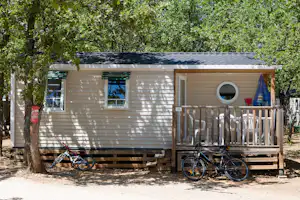 Mobilheim mit Holzterrasse, Fahrrädern davor und großen Fenstern, umgeben von Bäumen im Campingbereich.