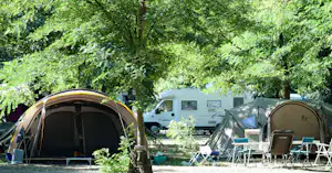Zelte und Wohnmobil auf dem Campingplatz im Schatten von Bäumen, mit Campingstühlen und Tisch am Stellplatz.