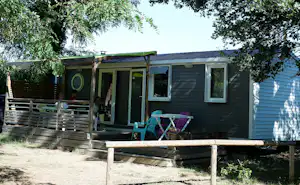 Mobilheim mit Terrasse, Holzzaun und Gartenmöbeln vor grünen Bäumen, mit Markise und Fenstern.