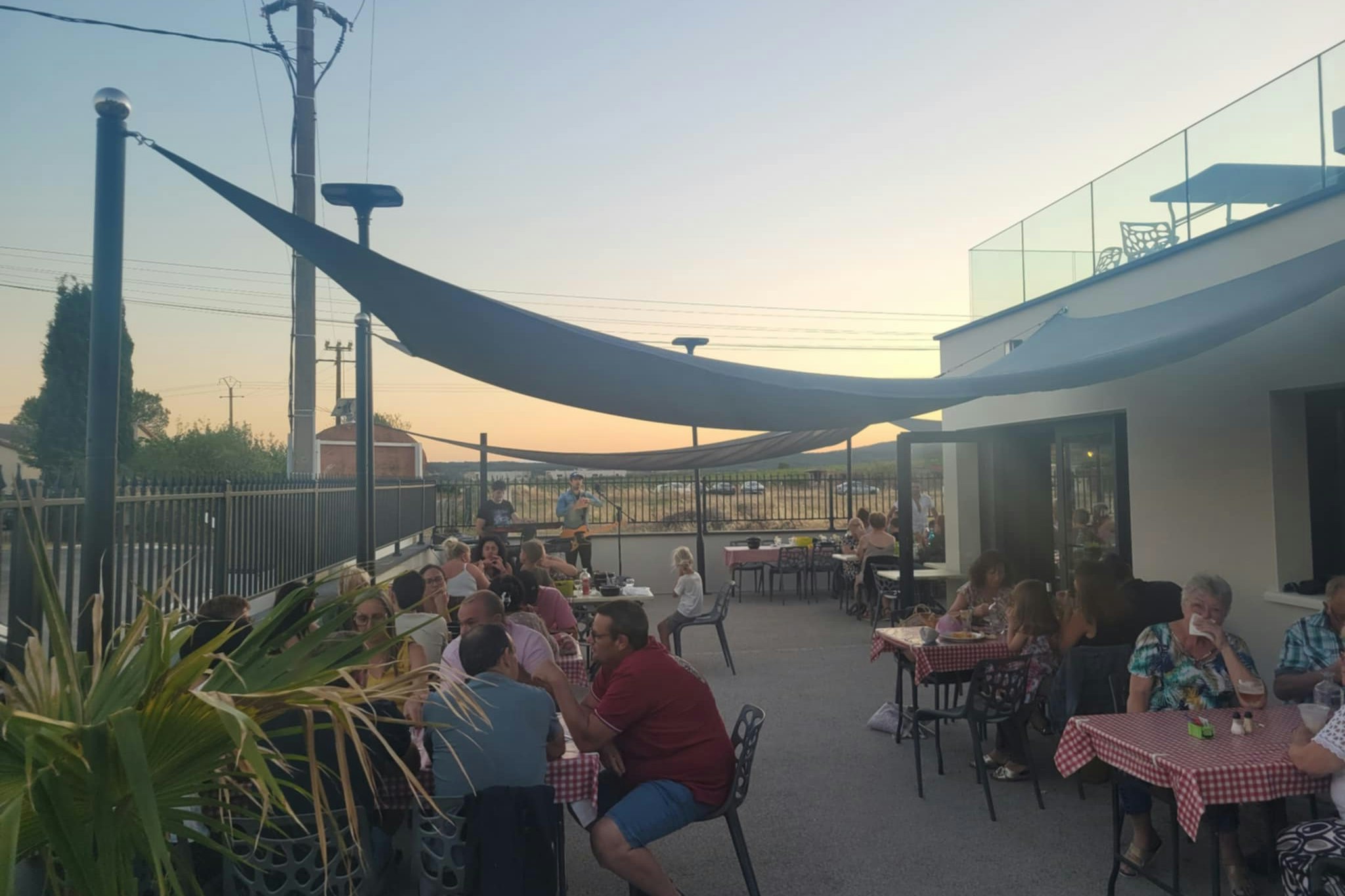 Camping-Terrasse mit Tischen und Stühlen unter großen Sonnensegeln, im Hintergrund Zaun und Gebäude bei Abendlicht.
