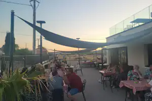 Camping-Terrasse mit Tischen und Stühlen unter großen Sonnensegeln, im Hintergrund Zaun und Gebäude bei Abendlicht.