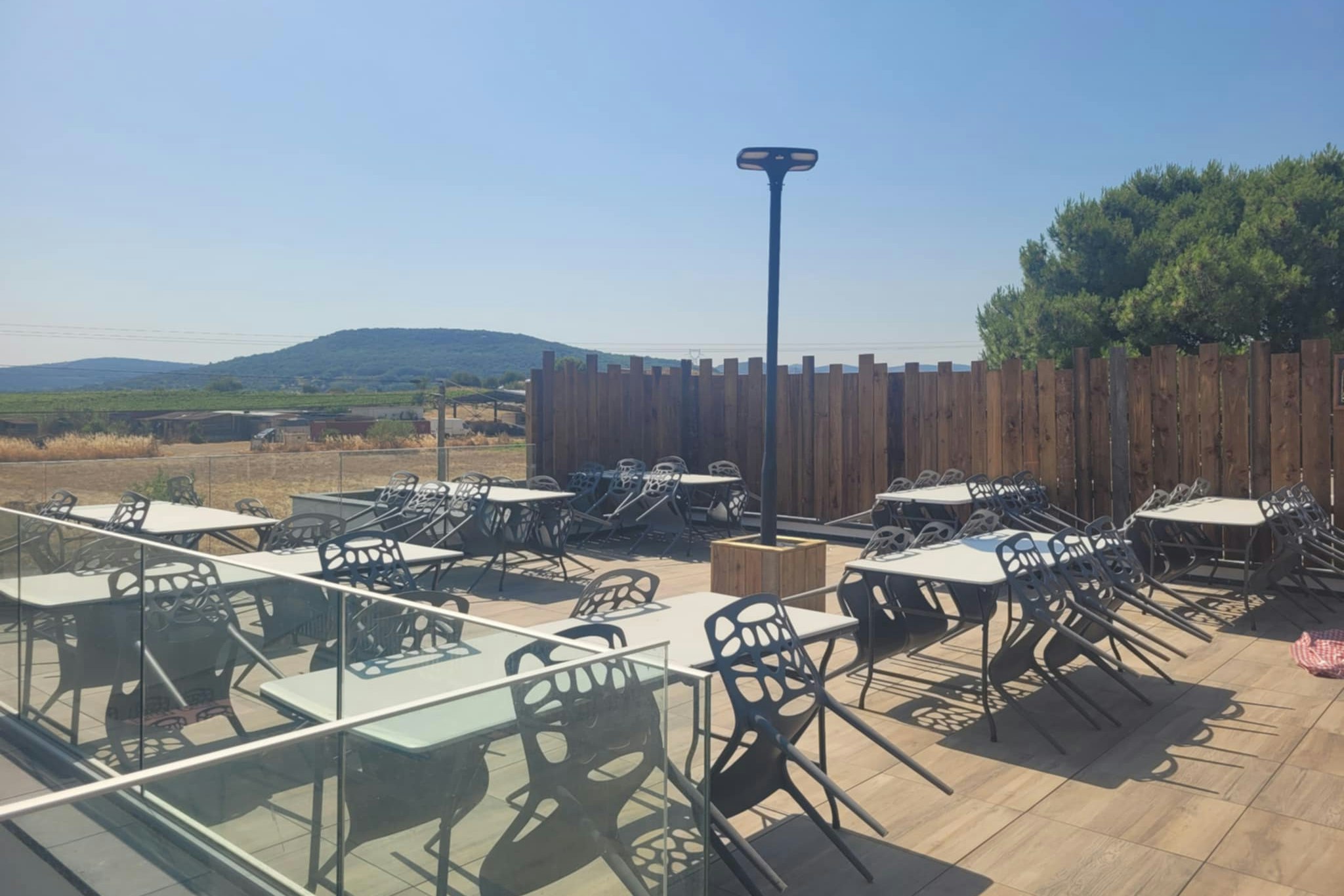 Terrasse mit vielen Tischen und Stühlen, Sonnenschirmständer und Holzzaun; im Hintergrund Wiesen und Hügel.