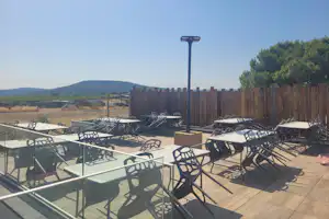 Terrasse mit vielen Tischen und Stühlen, Sonnenschirmständer und Holzzaun; im Hintergrund Wiesen und Hügel.