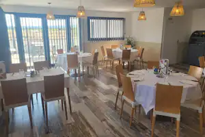 Essbereich im Campinghaus mit gedeckten Tischen, Holzstühlen, großen Fenstern und Vorhängen, daneben Grill auf der rechten Seite.