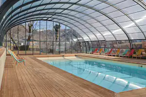 Beheiztes Schwimmbecken unter einem Glasdach mit Holzdeck und bunten Liegestühlen entlang der Seitenwand.