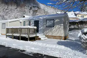 Mobilheim auf dem Campingplatz im Schnee, mit Holzveranda, großen Fenstern und verschneiten Bäumen im Hintergrund.