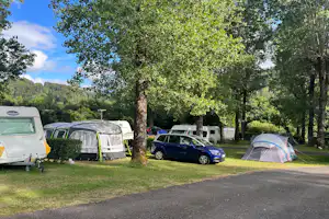 Campingplatz mit mehreren Zelten, Wohnwagen und geparkten Autos auf einer Rasenfläche zwischen Bäumen.