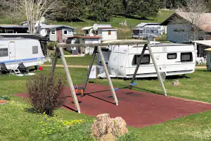 Wohnwagen und Camper auf einem Stellplatz, mit Holzschaukel auf roter Terrasse und Bäumen im Hintergrund.