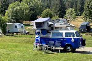 Blauer Camper-Van mit Stühlen und Leiter auf einem Campingplatz, daneben Zelt und Wohnwagen vor Waldkulisse.