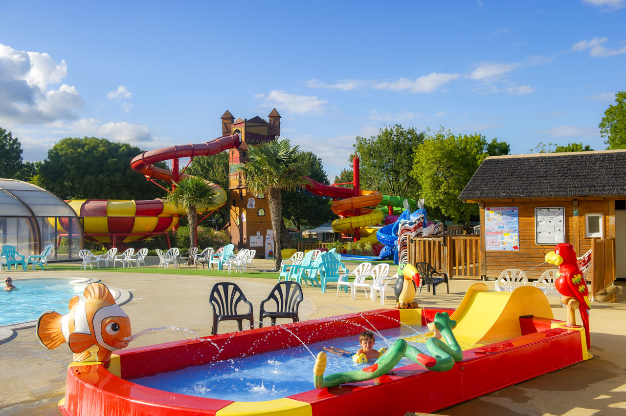 Wasserspiel im Campingpool mit roten Rutschen, bunten Liegestühlen und einem kleinen Holzhaus im Hintergrund.