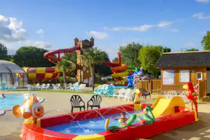 Wasserspiel im Campingpool mit roten Rutschen, bunten Liegestühlen und einem kleinen Holzhaus im Hintergrund.