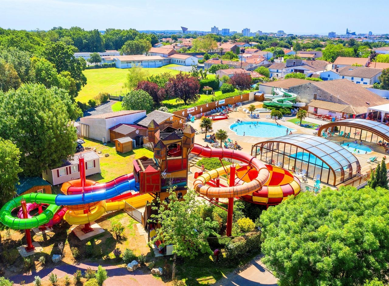 Luftaufnahme eines Campingplatzes mit Wasserpark, bunten Rutschen, beheiztem Pool und Gebäuden, umgeben von Bäumen und Grünflächen.