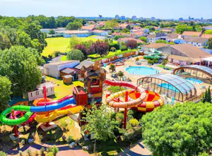 Luftaufnahme eines Campingplatzes mit Wasserpark, bunten Rutschen, beheiztem Pool und Gebäuden, umgeben von Bäumen und Grünflächen.
