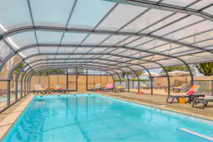 Beheiztes Hallenbad im Campingbereich mit überdachtem Glasdach, Liegen und Poolwasser, daneben gelbe Taschen und Zaun.