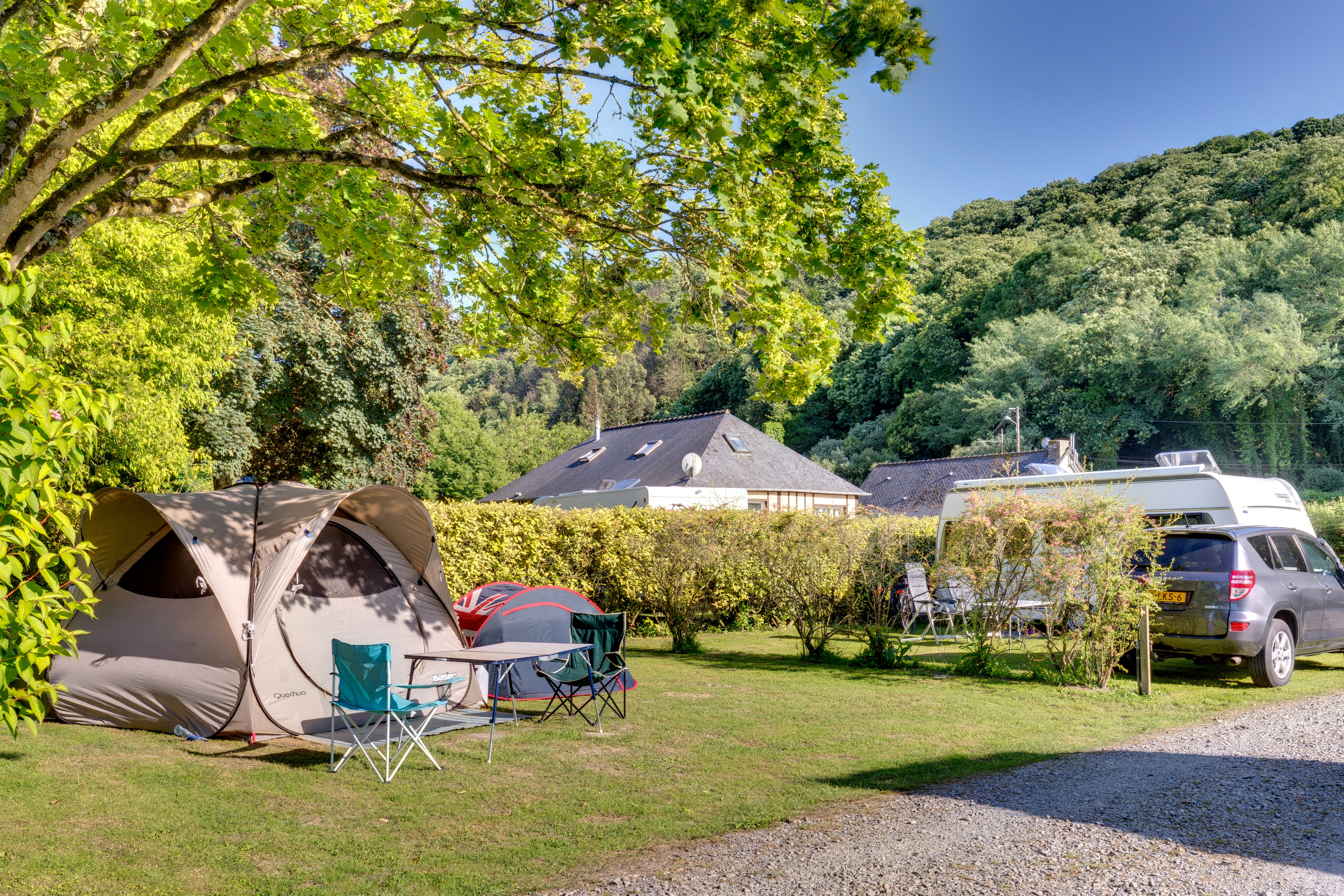 Campingzelt im Garten neben einem Stellplatz mit Wohnmobil und Auto, umgeben von Bäumen und Hecken.