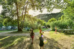 Kinder laufen über eine Wiese zwischen Bäumen, im Hintergrund Wohnwagen und ein Picknicktisch am Campingplatz.