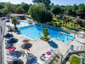 Großes Camping-Schwimmbecken mit Liegestühlen, Sonnenschirmen und gepflegtem Garten, daneben Gebäude und Wege im Hintergrund.