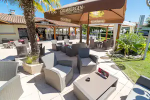 Terrasse mit Sitzgruppen aus grauen Polsterstühlen und Tischen, Sonnenschirmen, Palmen und gepflegtem Garten am Campingplatz.