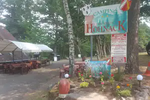 Campingplatz-Schild „Camping des Deux Hohnack“ am Weg, mit Bäumen, Sitzgarnitur und Zeltunterstand im Hintergrund.