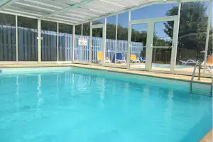 Überdachter Campingpool mit blauem Wasser, Glaswand und Liegestühlen im Hintergrund.