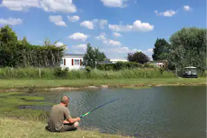 Angler sitzt am Teichufer und hält eine Angel, im Hintergrund Wohnwagen und Bäume auf dem Campingplatz.