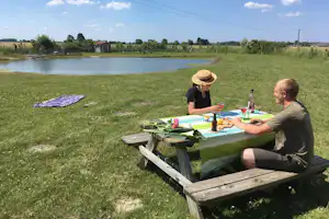 Camping-Tisch mit Getränken und Gemüse, zwei Personen sitzen am Ufer eines kleinen Sees im Grünen.