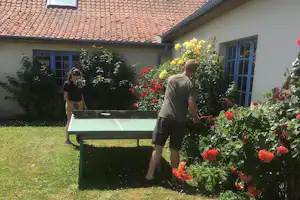 Tischtennisplatte vor einem weißen Gebäude, umgeben von roten und gelben Blumensträuchern und grünen Büschen.