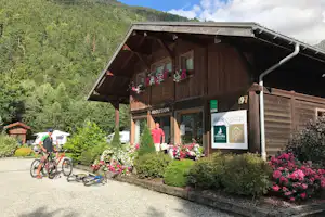 Rezeption im Holzgebäude „Nature Lodge“ mit Blumenbeeten, Fahrradstellplatz und Nummer 97 vor grünen Bergen.