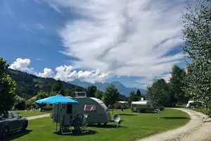 Wohnwagen mit Vorzelt und Sitzgruppe auf dem Campingplatz, Rasen und Bäume, im Hintergrund Berge und Wolken.