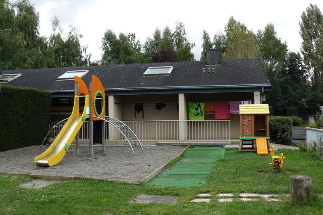 Spielplatz am Campingplatz mit gelber Rutsche, Kunstrasen und gepflegtem Garten vor einem Gebäude mit Terrasse.