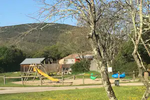 Spielplatz mit gelber Rutsche und Schaukel auf dem Campingplatz, im Hintergrund Gebäude und grüne Hügel.