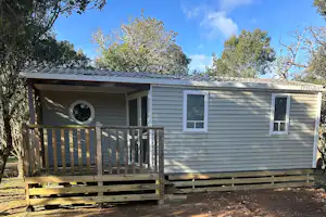Mobilheim mit Veranda und Holzgeländer, davor gepflasterter Stellplatz, umgeben von Bäumen und Schatten im Freien.