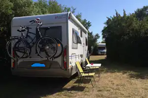 Wohnmobil mit Fahrrädern am Heck und Campingstuhl am Stellplatz, umgeben von Bäumen und Gras.