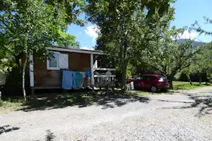 Holzbungalow auf dem Campingplatz mit Terrasse, Wäscheleine und rotem Auto, umgeben von Bäumen und Rasen.