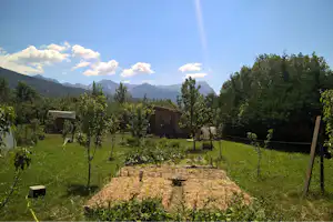 Campinggarten mit jungen Bäumen, Hochbeet und Holzschuppen, im Hintergrund Berge und grüner Wald unter blauem Himmel.