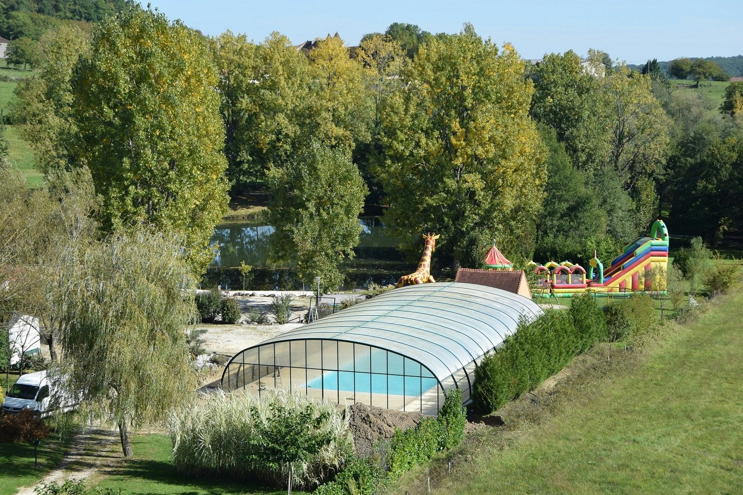 Überdachte Zeltkonstruktion auf einer Wiese, dahinter Bäume und ein Spielplatz mit Rutsche und bunten Elementen.
