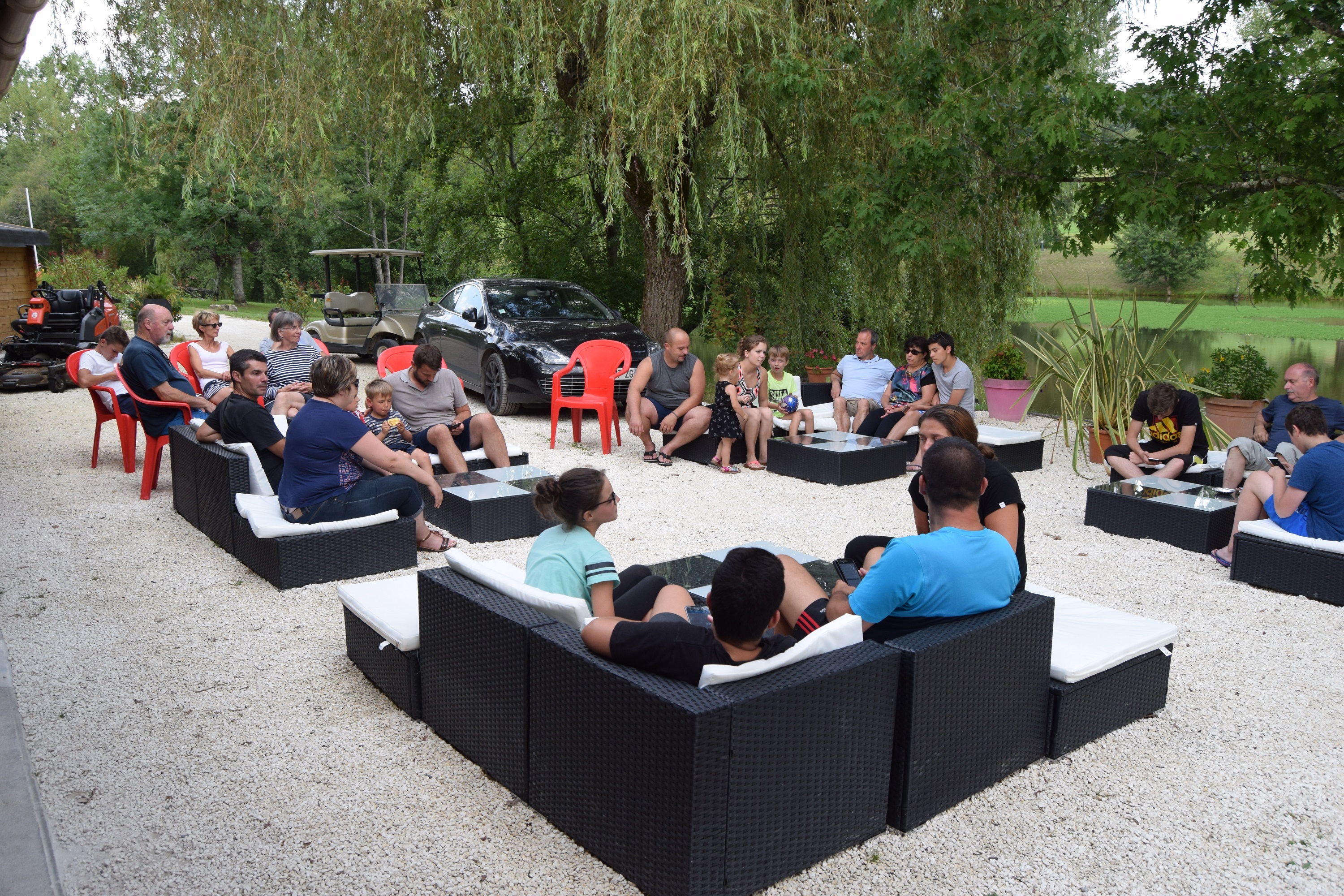 Gruppe sitzt auf einer Camping-Terrasse in schwarzen Lounge-Sesseln, daneben Tische und Stühle, im Hintergrund Autos und Bäume.