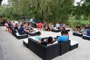 Gruppe sitzt auf einer Camping-Terrasse in schwarzen Lounge-Sesseln, daneben Tische und Stühle, im Hintergrund Autos und Bäume.