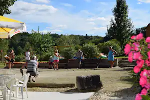 Boulespiel auf einem Campingplatz mit Rasenfläche, Holzbank und Bäumen im Hintergrund, im Vordergrund Blumen und Sonnenschirm.