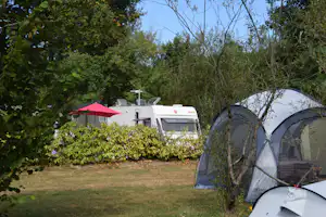 Campingzelt am Rand eines Rasens, im Hintergrund Wohnwagen mit rotem Sonnenschirm und Hecke.