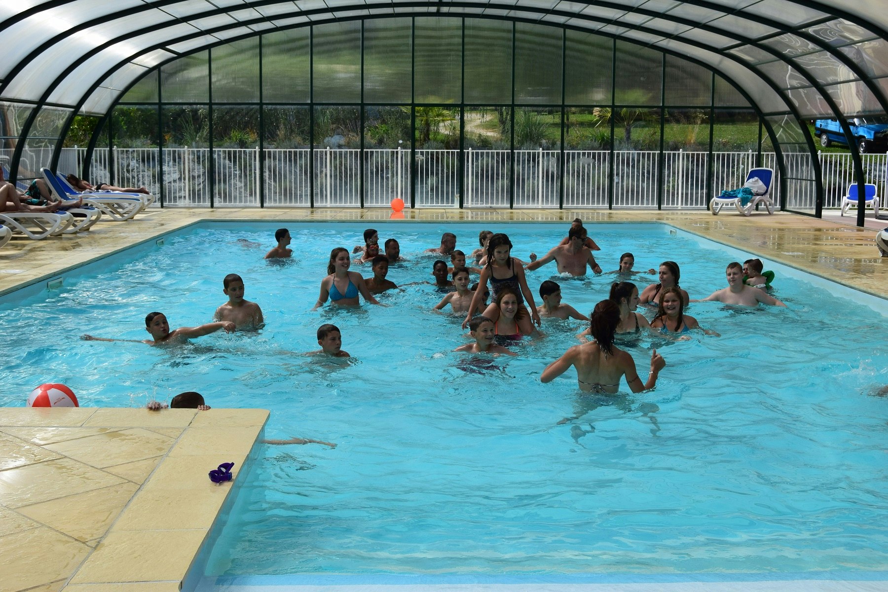 Beheiztes Schwimmbecken im Campingbereich mit vielen Badegästen, Glasdach und Liegen am Beckenrand.