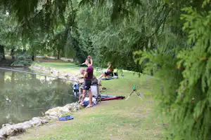 Campingplatz im Grünen: Person am Grill neben Zelt und Campingstühlen auf einer Wiese, im Hintergrund Bäume und Hecke.