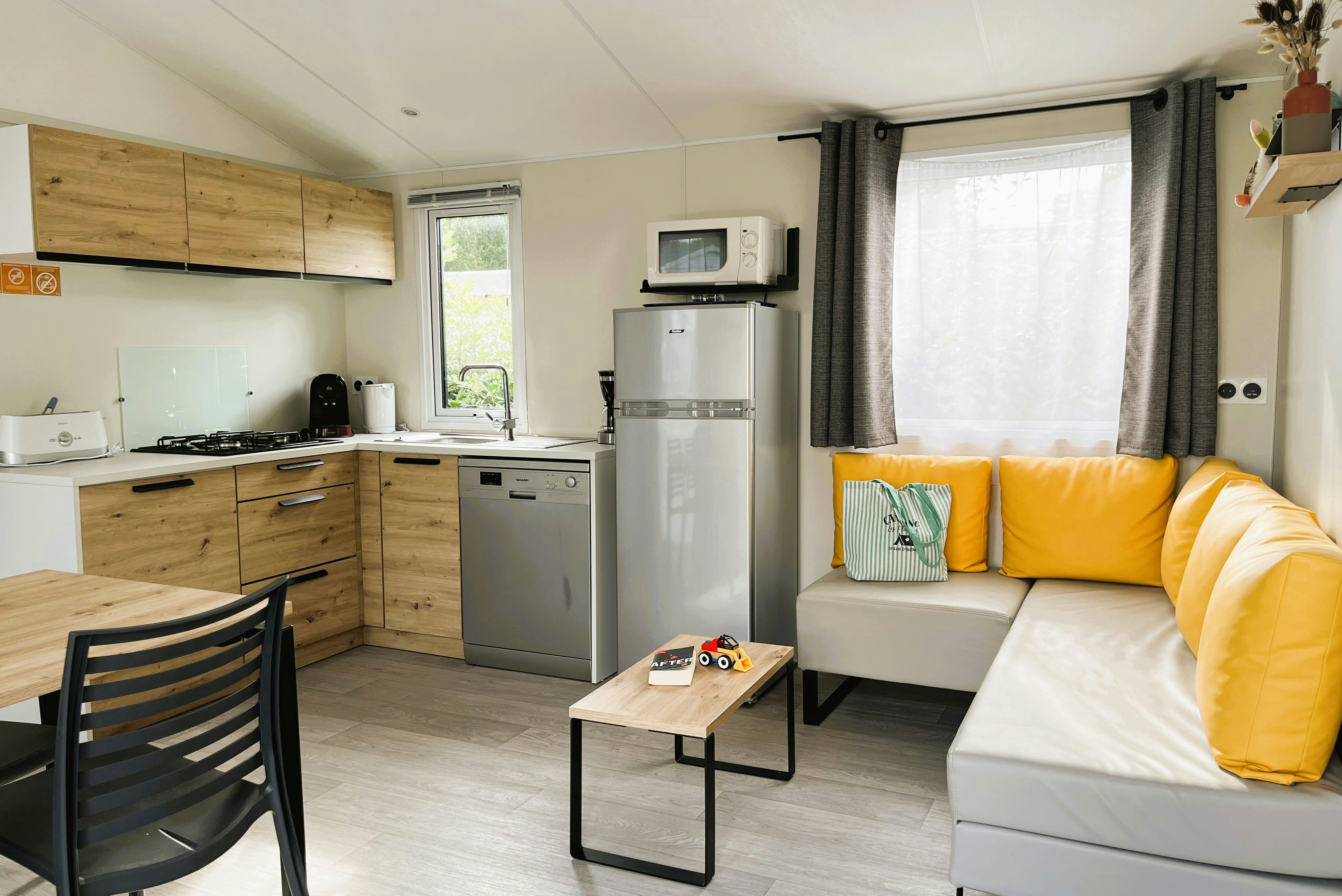 Wohnbereich mit Küchenzeile, Sofa und Couchtisch in einem Camping-Mobilheim, mit Fenster und Vorhängen.