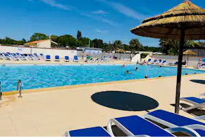 Blauer Pool mit Liegestühlen und Sonnenschirmen am Campingplatz, im Hintergrund Gebäude und Bäume unter klarem Himmel.