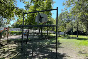 Spielplatz mit eingezäuntem Klettergerüst auf einem Campingplatz, umgeben von Bäumen und Rasenfläche.