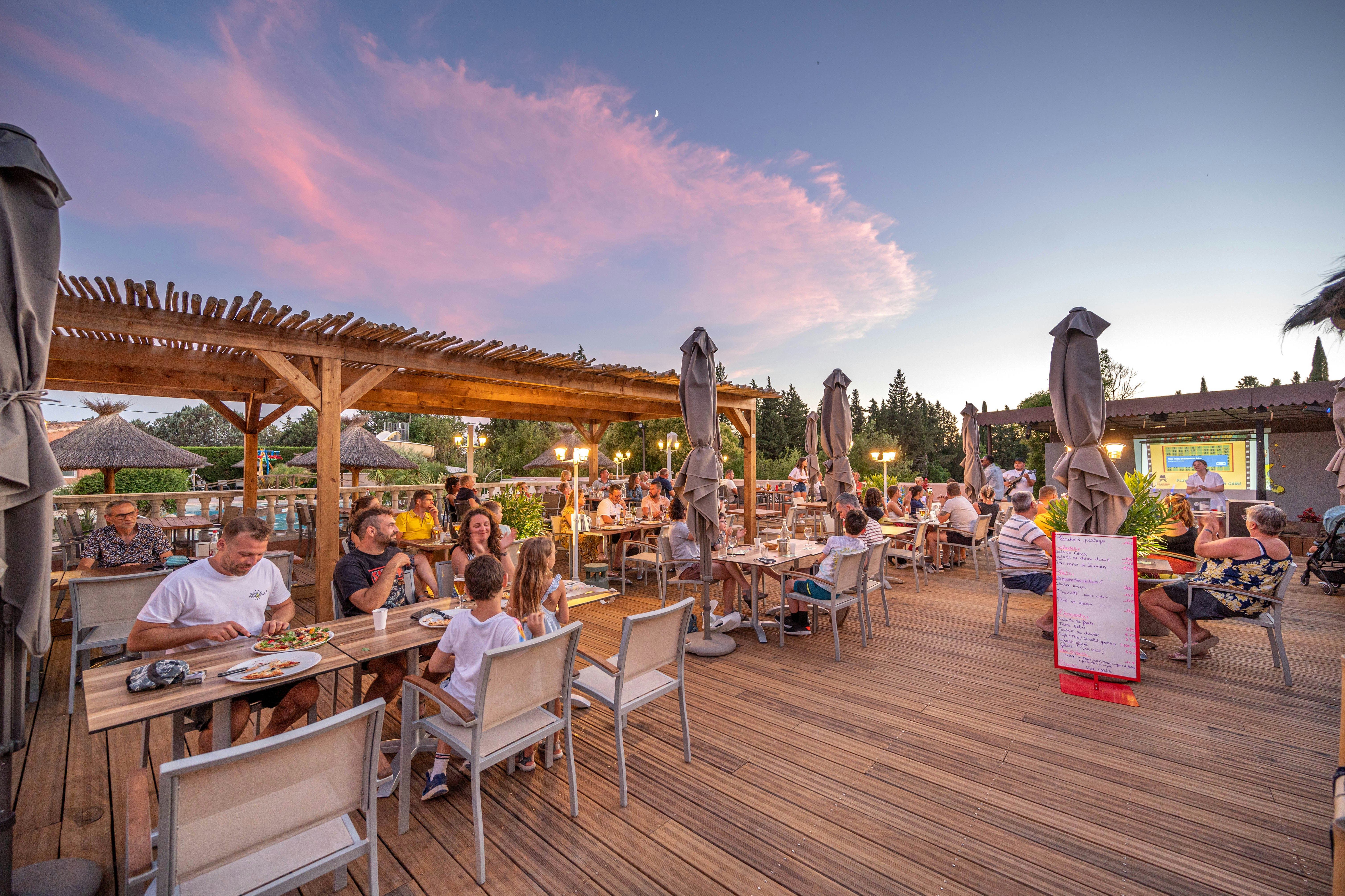 Holzterrasse eines Campingplatzes mit gedeckten Tischen, Stühlen und Sonnenschirmen, Gäste sitzen am Abend.