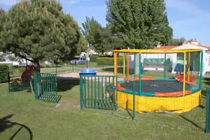 Gelbes Trampolin in einem eingezäunten Spielbereich auf dem Campingplatz, mit Rasen, Bäumen und bunten Spielgeräten im Hintergrund.